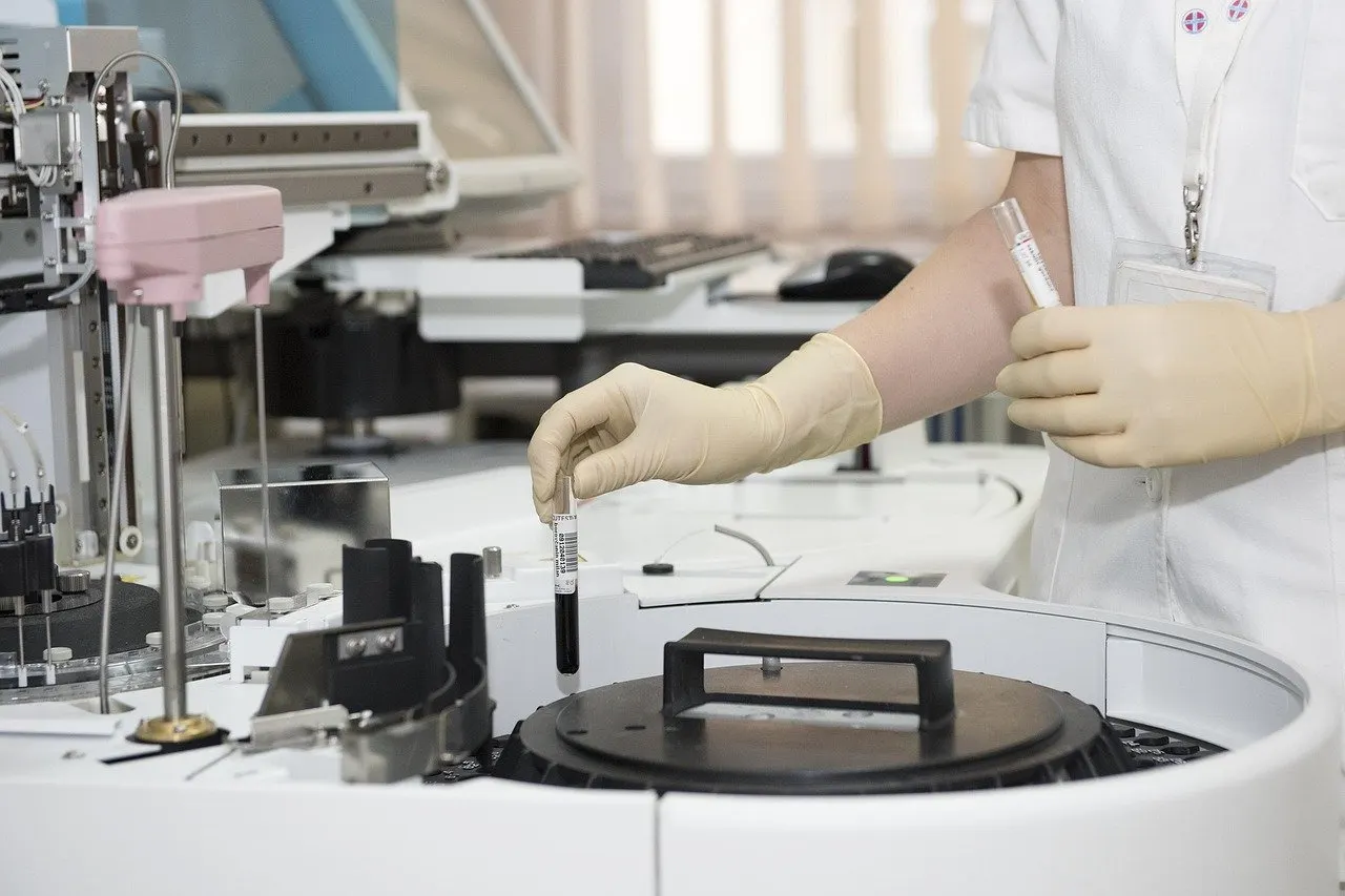 Hände in Laborhandschuhen setzen ein Blutröhrchen in ein Analysegerät, ein zweites Röhrchen in der anderen Hand, im Hintergrund Tastatur und Laborgeräte