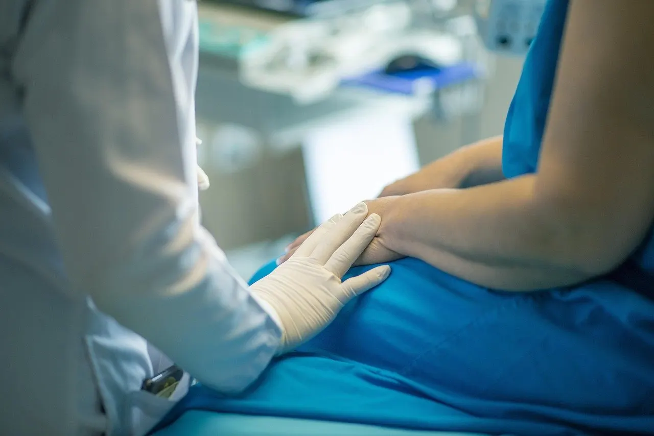 Behandelnde Person im weißen Kittel mit Handschuhen legt die Hand beruhigend auf die Hand einer Patientin in blauem Untersuchungshemd auf der Liege
