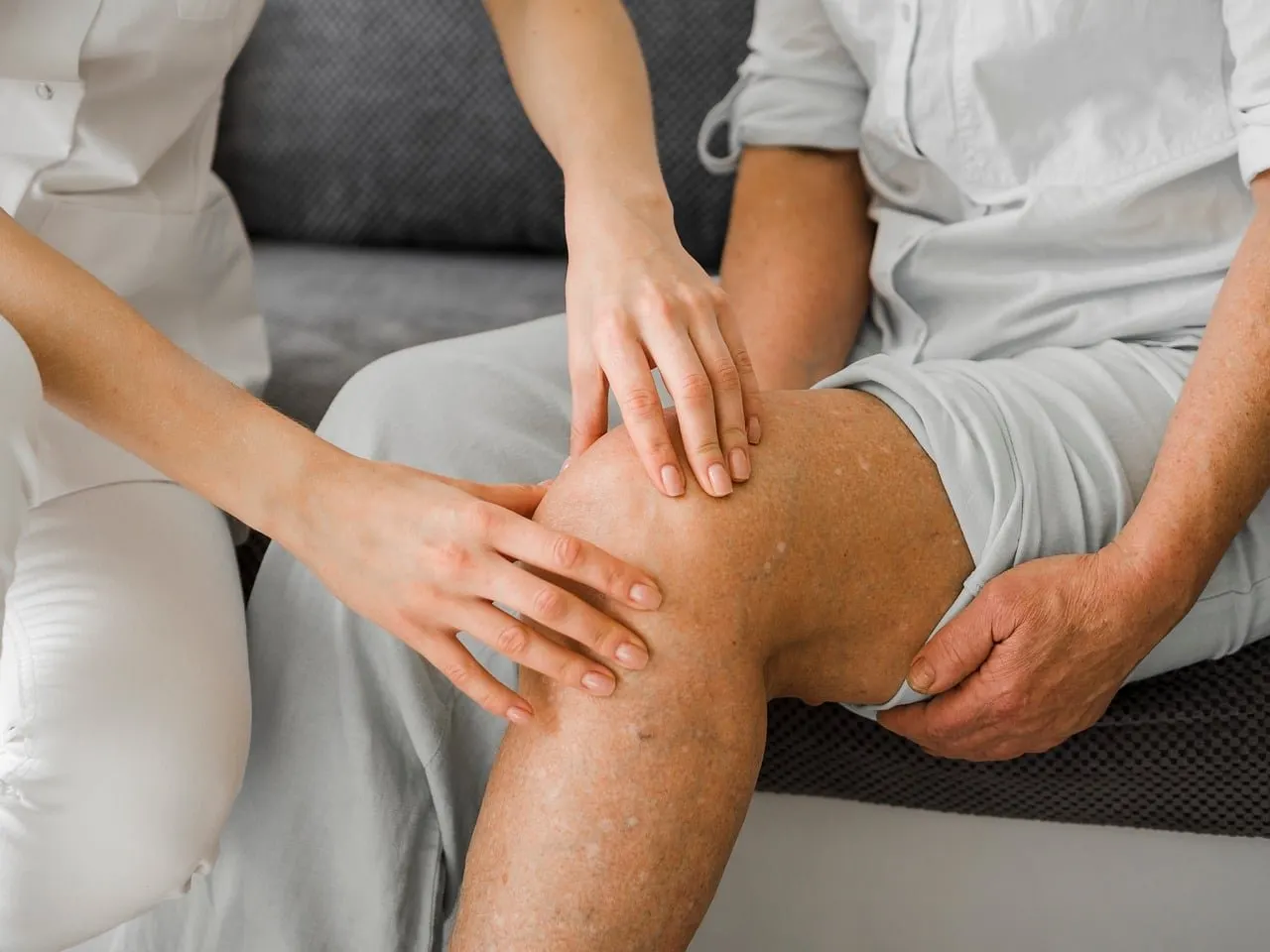 Hände einer Therapeutin tasten das Knie einer sitzenden älteren Person ab, die sich mit der Hand am Oberschenkel hält
